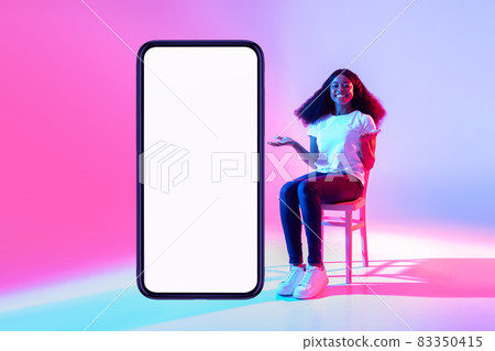 Lovely black lady sitting on chair near modern smartphone with empty screen and offering space for mockup in neon light Lovely black lady sitting on chair near modern smartphone with empty screen and offering space for mockup in neon light 83350415