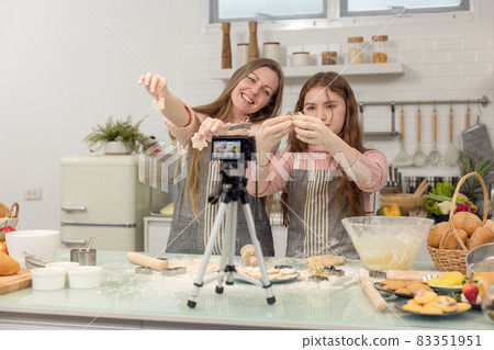 With a live digital camera in the kitchen, mother and daughter smile and have fun while preparing biscuit dough together, daughter learning how to make cookies with her mother. 83351951
