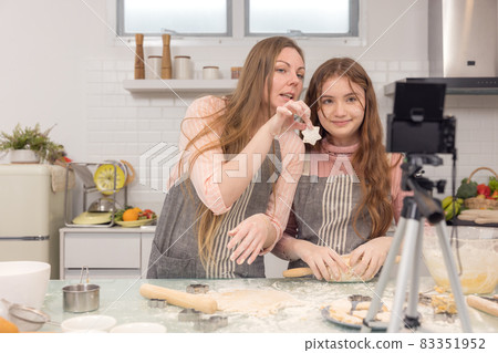 With a live digital camera in the kitchen, mother and daughter smile and have fun while preparing biscuit dough together, daughter learning how to make cookies with her mother. 83351952