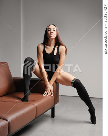 Middle age woman sits on sofa in black bodysuit and warm overknee socks 83353427