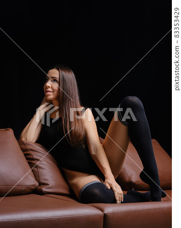 Middle age woman sits on sofa in black bodysuit and warm overknee socks 83353429