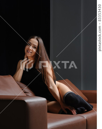 Middle age woman in bodysuit and overknee socks sits on leather sofa 83353430