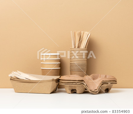 stack of brown disposable paper cups and a tray on a white table, wooden forks and knives, brown background stack of brown disposable paper cups and a tray on a white table, wooden forks and knives, brown background 83354903
