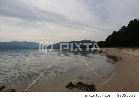 Rippled sea surface, sandy beach with large curved stones, pine forest, distant city, mountains and cloudy sky Rippled sea surface, sandy beach with large curved stones, pine forest, distant city, mountains and cloudy sky 83355558