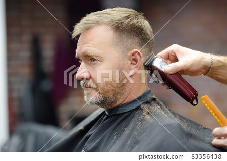 Barber master is shaving of handsome mature bearded man in salon. Hair artist making hairstyle for person in barbershop. Services of professional stylist. 83356438