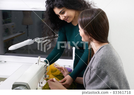 Pretty seamstress teaching girl working with sewing machine at sewing class in tailor studio Pretty seamstress teaching girl working with sewing machine at sewing class in tailor studio 83361718