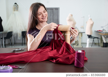 Beautiful young dressmaker in workroom sewing red dress in tailor studio or atelier shop Beautiful young dressmaker in workroom sewing red dress in tailor studio or atelier shop 83366880