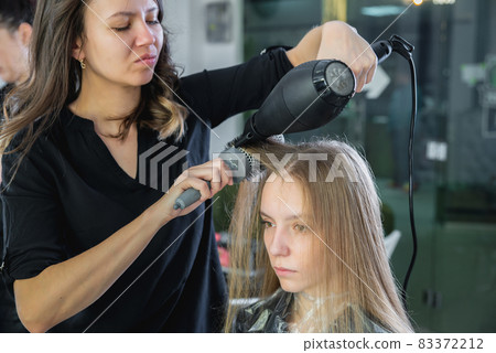 Close up of hairdressers hands drying long blond hair with blow dryer and round brush 83372212