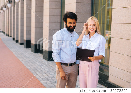 Medium shot portrait of cheerful male and female colleagues discussing document on clipboard standing in city street by office building. Medium shot portrait of cheerful male and female colleagues discussing document on clipboard standing in city street by office building. 83372831