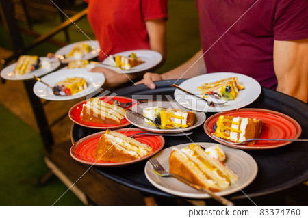 Close-up of the waiter's girl's hands holding white plates with cut pieces of cake. The waiter serves desserts. 83374760
