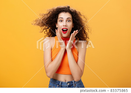 Waist-up shot of charmed enthusiastic attractive female with curly hairstyle in cropped top holding palms on cheeks and smiling astonished and surprised gazing with amazement over orange background Waist-up shot of charmed enthusiastic attractive female with curly hairstyle in cropped top holding palms on cheeks and smiling astonished and surprised gazing with amazement over orange background 83375865