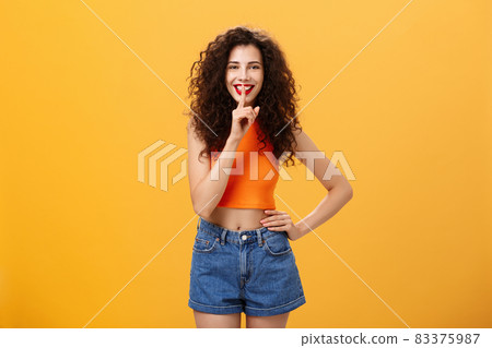 Shh I got something for you. Portrait of charming european young female with curly hairstyle in cropped top and shorts showing shush gesture with broad smile having secret preparing surprise Shh I got something for you. Portrait of charming european young female with curly hairstyle in cropped top and shorts showing shush gesture with broad smile having secret preparing surprise 83375987