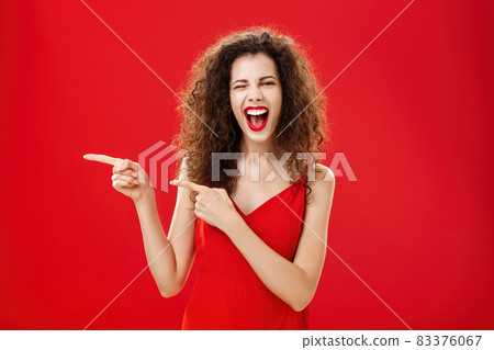 Woman feeling awesome on great party of friend pointing at entrance. Stylish carefree curly-haired female in red evening dress having fun near pool winking smiling joyfully and pointing left 83376067