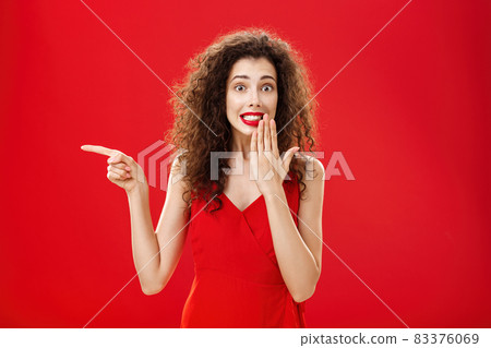 Troubled cute and tender silly woman with curly hairstyle in red stylish evening dress saying oops smiling with guilty look covering mouth with palm while pointing left over studio background 83376069