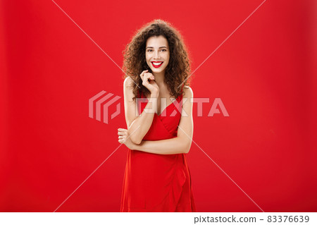Passionate charming european woman over red background in elegant dress with curly hairstyle smiling cute, feminine playing with hair strand standing timid and silly, talking to person she admires Passionate charming european woman over red background in elegant dress with curly hairstyle smiling cute, feminine playing with hair strand standing timid and silly, talking to person she admires 83376639