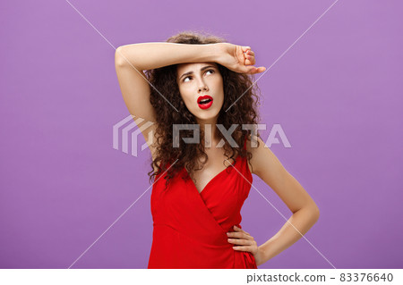 Troubled and tired good-looking european woman with curly hairstyle in red evening dress wiping sweat of forehead looking at upper right corner perplexed and exhausted over purple background 83376640