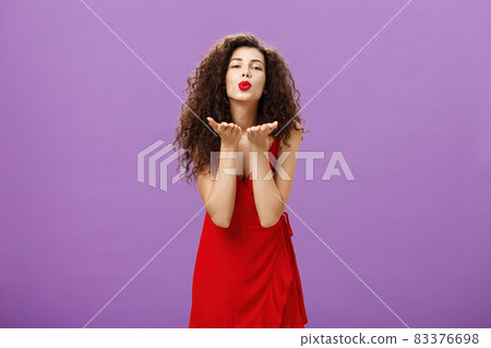 Tender charming european woman with red lipstick in elegant evening dress bending towards camera folding lips sending air kiss at camera holding palms near mouth expressing love and passion Tender charming european woman with red lipstick in elegant evening dress bending towards camera folding lips sending air kiss at camera holding palms near mouth expressing love and passion 83376698