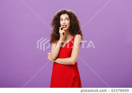 Silly and timid charming woman with dark curly hair in stylish evening dress with red lipstick biting finger looking cute at upper right corner thoughtful imaging or dreaming about desirable thing 83376699