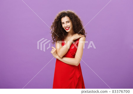 Shy feminine elegant european female with curly hairstyle and lipstick in red luxurious dress touching shoulder gently chuckling over funny jokes of boyfriend during romantic dinner over purple wall 83376700