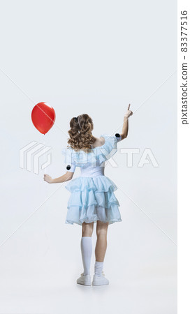 Back view of young girl wearing Halloween dress, costume of movie character standing with red balloon in hand isolated over blue background Back view of young girl wearing Halloween dress, costume of movie character standing with red balloon in hand isolated over blue background 83377516