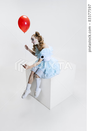 Top view of young girl wearing Halloween dress of dancing clown with spooky make-up sitting on box with red balloon 83377597