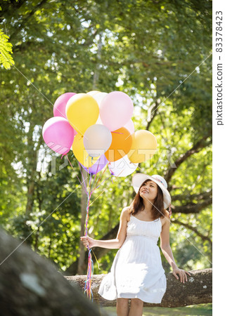 Cheerful beauty woman holding balloons relax sitting under big tree in green park with happiness. Woman Hands holding vibrant air balloons play on birthday party happy time summer on sunshine outdoor 83378423
