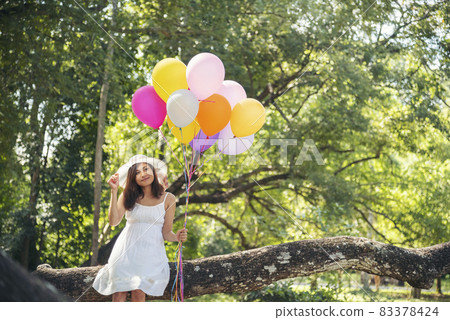 Cheerful beauty woman holding balloons relax sitting under big tree in green park with happiness. Woman Hands holding vibrant air balloons play on birthday party happy time summer on sunshine outdoor 83378424