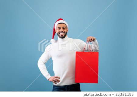 Happy arab man wearing Santa hat and holding shopping bag with Christmas gift over blue background, studio shot Happy arab man wearing Santa hat and holding shopping bag with Christmas gift over blue background, studio shot 83381193