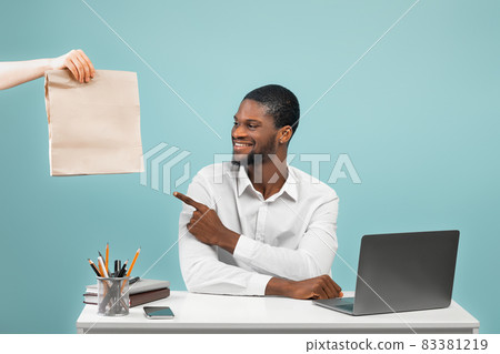 African american company worker receiving food delivery from courier and pointing on paper bag, blue background African american company worker receiving food delivery from courier and pointing on paper bag, blue background 83381219