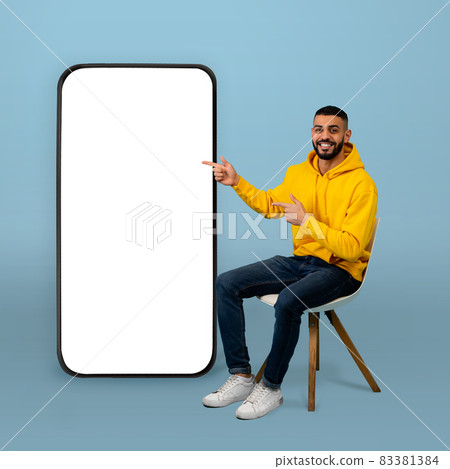 Positive arab guy pointing at big smartphone with white screen while sitting on chair over blue background Positive arab guy pointing at big smartphone with white screen while sitting on chair over blue background 83381384