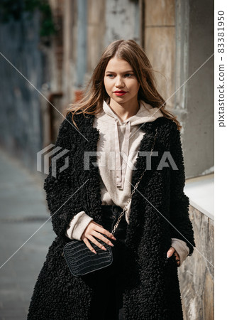 Close-up photo of young beautiful woman in black fur coat, jeanse and shoes posing on street Close-up photo of young beautiful woman in black fur coat, jeanse and shoes posing on street 83381950