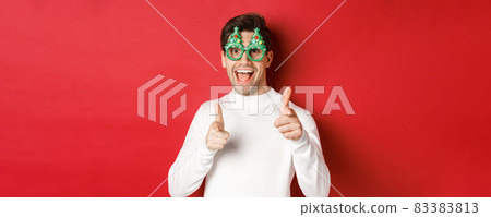 Joyful caucasian guy in party glasses and white sweater, smiling and pointing fingers at camera, wishing merry christmas and happy new year, standing over red background 83383813
