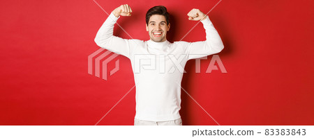 Portrait of smiling handsome man in white sweater, flexing biceps and bragging with strength, show-off strong muscles after workout, standing over red background 83383843