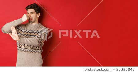 Image of disgusted handsome man in winter sweater, rejecting something with bad smell, shut nose and raising hand to decline, standing over red background 83383845