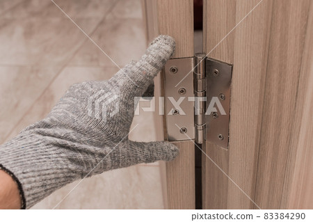 The hand of a working man in a construction glove touches and examines a wooden door opening with a door hinge, close-up. Installation or adjustment of metal parts 83384290