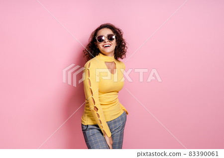 Laughing curly girl dressed yellow outfit and sunglasses while posing isolated over pink background. 83390061
