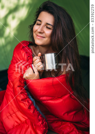 A happy tourist woman is sitting in a tent and drinking tea. Camp in the tent - young girl on the camping. A female tourist in a sleeping bag, drinking hot tea. Morning at the campsite. Portrait A happy tourist woman is sitting in a tent and drinking tea. Camp in the tent - young girl on the camping. A female tourist in a sleeping bag, drinking hot tea. Morning at the campsite. Portrait 83392259
