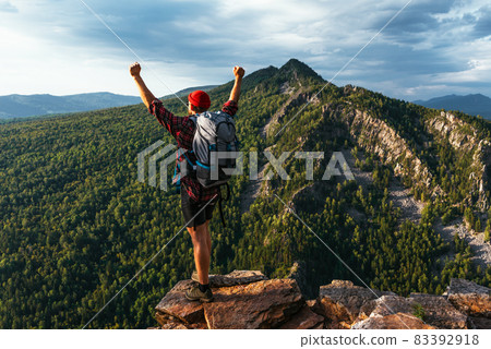 Hiker with arms up standing on the top of the mountain. A man with a backpack in the mountains at sunset, rear view. A trip to the mountains with a backpack. A traveler on the background of mountains 83392918