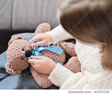 little girl playing with knitted teddy bear and ties a ribbon or bow. Top view, unrecognizable person. knitting as hobby, hand made toys little girl playing with knitted teddy bear and ties a ribbon or bow. Top view, unrecognizable person. knitting as hobby, hand made toys 83393140