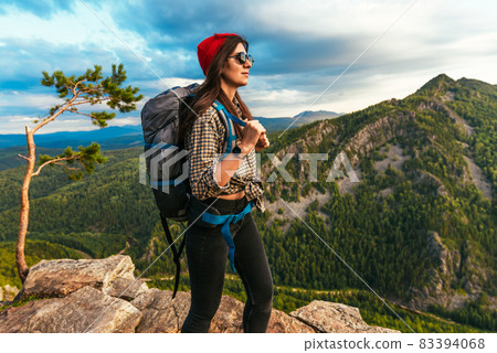 A young tourist girl with a backpack enjoys the sunset from the top of the mountain. Portrait of a traveler in a red riding hood. A tourist with a backpack is standing on the top of the mountain 83394068