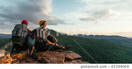 The couple meets the sunset in the mountains. Two travelers are sitting on the edge of a cliff and admiring the beautiful panoramic view. Travelers with backpacks at sunset in the mountains. Panorama 83394069