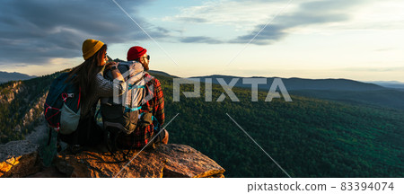 The couple meets the sunset in the mountains. Two travelers are sitting on the edge of a cliff and admiring the beautiful panoramic view. Travelers with backpacks at sunset in the mountains. Panorama 83394074