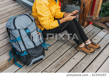 A traveler with a phone, close-up. A tourist girl in a yellow jacket with a backpack holds a phone in her hands. Hiking woman using smart phone, travel and active lifestyle concept. Copy space 83395097