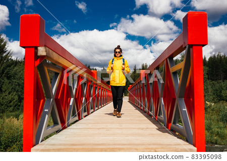 A young tourist girl in a yellow jacket with a backpack is walking along the red bridge. A tourist walks through the national park. A tourist passes the river through the bridge. Copy space 83395098