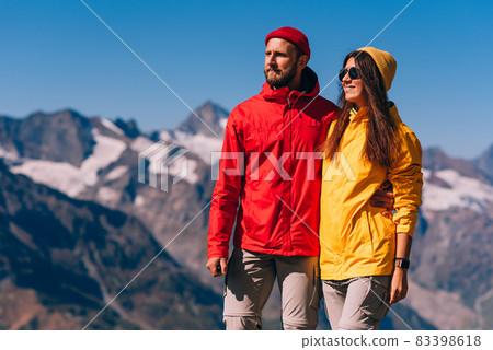 A traveling couple in hiking gear against the background of mountains. Two tourists on the top of the mountain. A man and a woman in the mountains. Two travelers in colorful jackets, close-up. 83398618