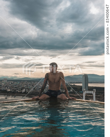 The view from the hotel rooftop pool of the city and the guy sitting on the edge of the rooftop swimming pool. Picturesque view, beautiful sky The view from the hotel rooftop pool of the city and the guy sitting on the edge of the rooftop swimming pool. Picturesque view, beautiful sky 83400647