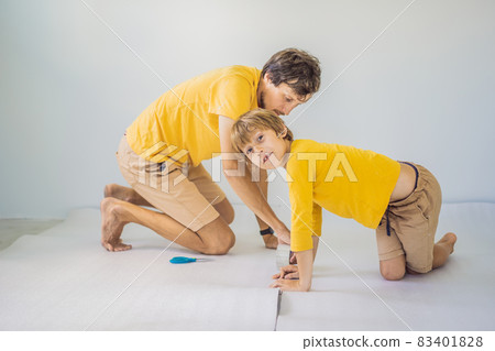 Dad and son are laying floor insulation. Roll of insulating cloth to install a laminate wooden floor Dad and son are laying floor insulation. Roll of insulating cloth to install a laminate wooden floor 83401828