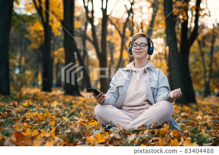 Caucasian middle aged woman in earphones listening to music, meditation app on smartphone and meditating in lotus pose at autumn park. Meditation app, Mental health, self care, mindfulness concept 83404488