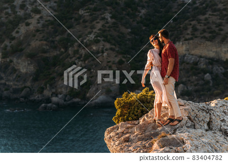 A man embraces a beautiful woman on the edge of a cliff by the sea. A couple in love at sunset. Beautiful couple meets sunset by the sea. Happy couple having fun on the coast. Copy space A man embraces a beautiful woman on the edge of a cliff by the sea. A couple in love at sunset. Beautiful couple meets sunset by the sea. Happy couple having fun on the coast. Copy space 83404782