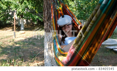 Female wearing virtual reality glasses rests in a hammock near a tree, imagining a dream come true 83405545